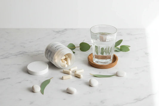 Magnesium Dosierung im Alltag: Eine umgekippte weiße Dose, aus der Magnesium Kapseln auf eine helle Marmorfläche rollen, daneben ein Glas Wasser zur Einnahme sowie Eukalyptusblätter.